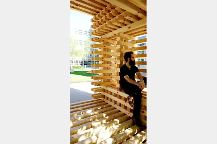 Person seated within a stacked wooden lattice pavilion casting striped shadows on the floor, with a sunlit lawn and buildings in the background. Person seated within a stacked wooden lattice pavilion casting striped shadows on the floor, with a sunlit lawn and buildings in the background.