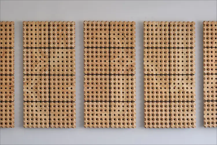 Wall-mounted panels of perforated wood tiles arranged in vertical grids with varied hole patterns on a white wall. Wall-mounted panels of perforated wood tiles arranged in vertical grids with varied hole patterns on a white wall.
