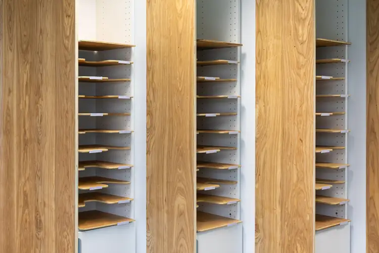 Vertical wooden shelving units with staggered trays set between tall wood panels, forming a repeating storage display against a neutral wall. Vertical wooden shelving units with staggered trays set between tall wood panels, forming a repeating storage display against a neutral wall.