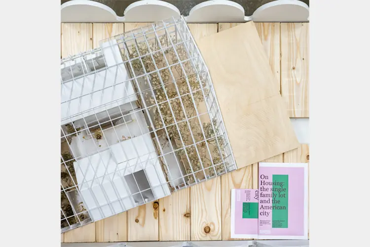 Architectural model in a wire frame enclosure on a wood surface, with plant material, plywood panels, and a housing-related booklet. Architectural model in a wire frame enclosure on a wood surface, with plant material, plywood panels, and a housing-related booklet.