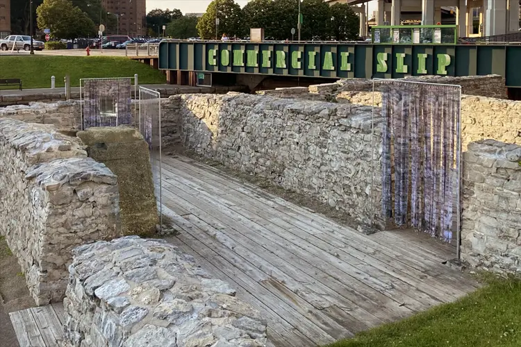 Stone foundation walls and wooden walkway at a historic archaeological site beneath a bridge sign reading “Commercial Slip.” Stone foundation walls and wooden walkway at a historic archaeological site beneath a bridge sign reading “Commercial Slip.”