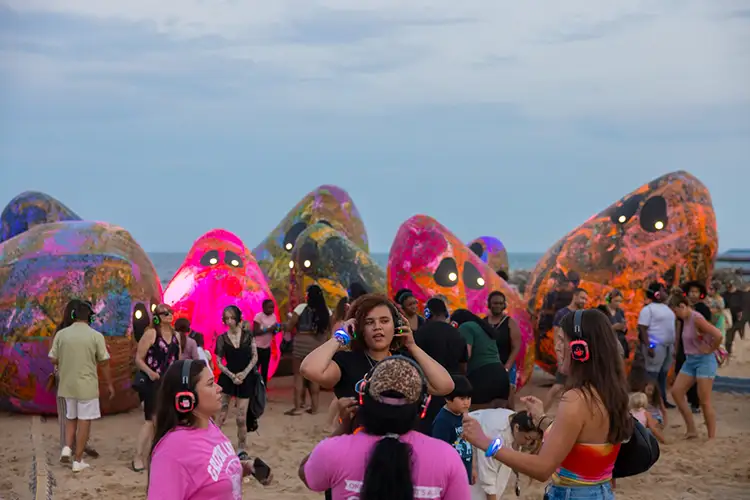 People wear headphones on a beach with large colorful inflatable figures behind them.