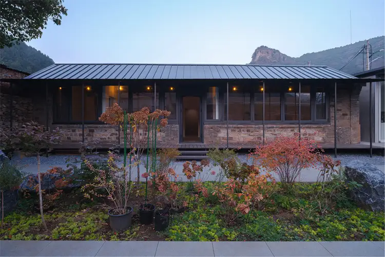 Single-story brick-and-stone building with a dark metal roof, warm interior lights, and a landscaped courtyard backed by wooded hills.
