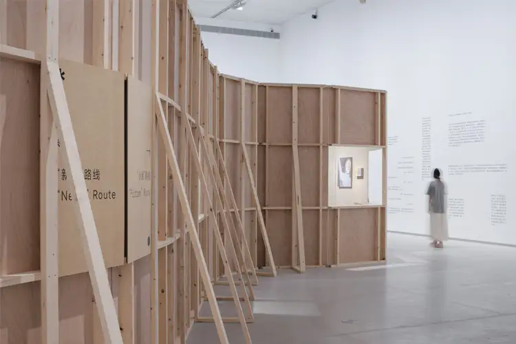 Curved plywood exhibition wall with exposed framing in a white gallery, featuring display niches, wall text, and a visitor walking nearby.