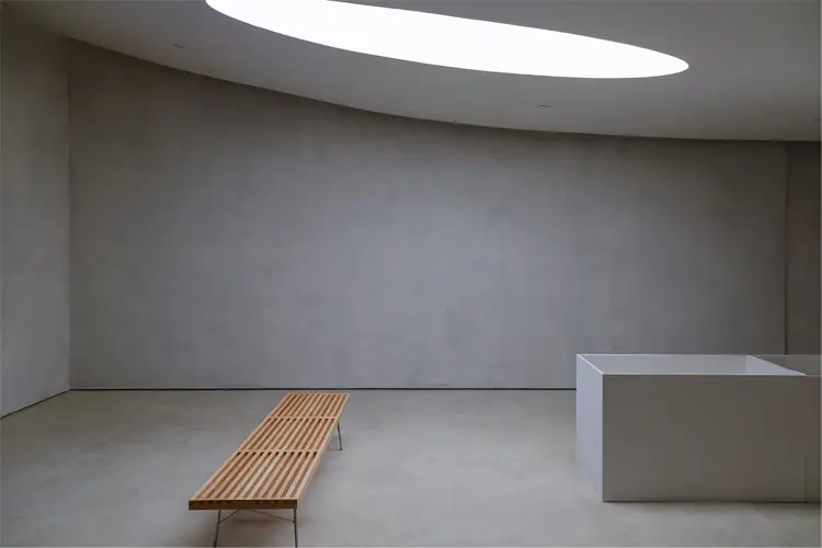 Minimalist gallery interior with a curved concrete wall, long wooden bench, white display plinth, and oval skylight overhead.