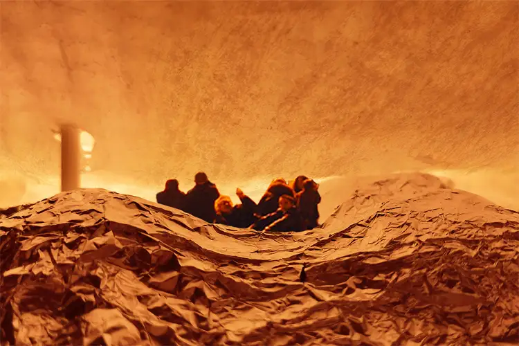 Visitors sit among crumpled paper-like sculpted forms in a warm, amber-lit installation that resembles a landscape of rolling hills.