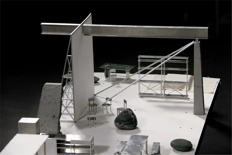 Architectural model with steel beams, angled trusses, seating, tables, and stone elements arranged on a white display platform.