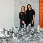 Two people stand behind a detailed architectural city model displayed in a design studio.