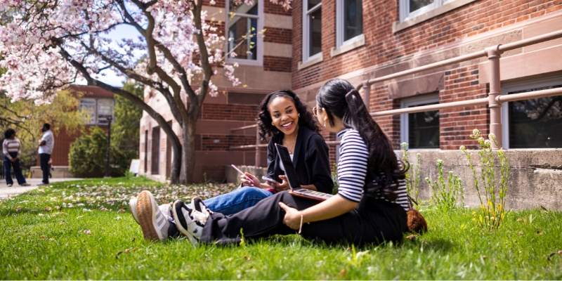Two students talking on the grass and working on homework outside of Mitchell Hall