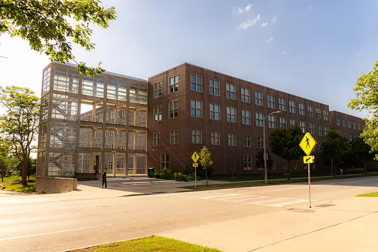 Red brick campus building at sunset with lit windows, glass walkway, and pedestrian near street crossing.