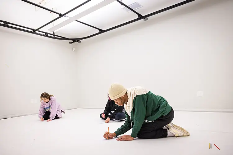 Three people kneel on a large white floor, drawing in a studio space.