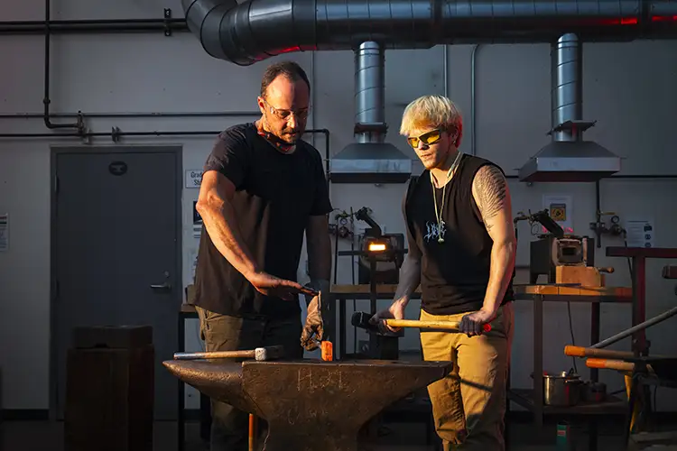 Two people working together at an anvil in a metal workshop.