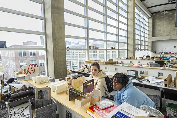 Two architecture students working on building models in a bright studio with large windows