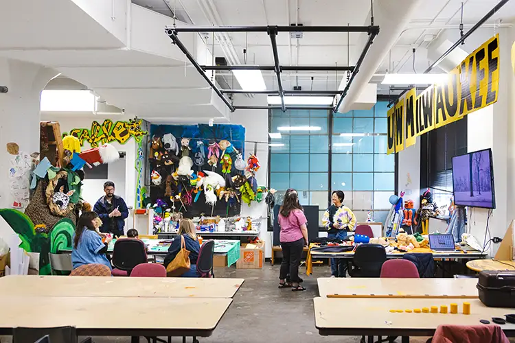 People working in a bright studio filled with art supplies, masks, and creative projects.