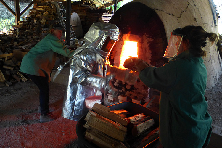 Two people wearing protective gear put material into a glowing wood fire kiln.