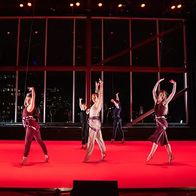 A group of dancers perform on a bright red stage, each with one arm raised. The city at night can be seen through large windows behind them.