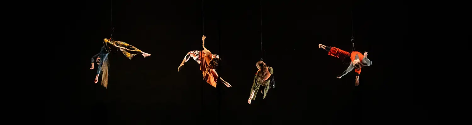 Four aerial performers in colorful costumes suspended midair against a black background
