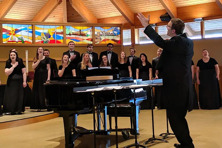 Choir performs beside a grand piano as a conductor leads with raised arms beneath stained-glass windows. Choir performs beside a grand piano as a conductor leads with raised arms beneath stained-glass windows.