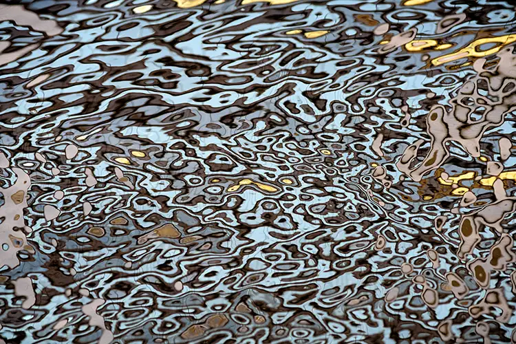 Abstract close‑up of rippling water creating swirling, mosaic‑like reflections in blue, black, and gold tones.