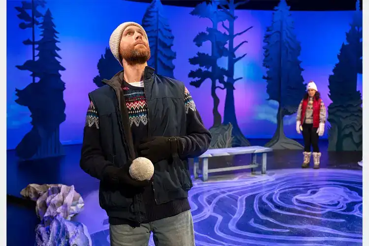 Performer in winter attire holds a snowball on a stage with stylized trees, while another bundled performer stands near a bench in the background. Performer in winter attire holds a snowball on a stage with stylized trees, while another bundled performer stands near a bench in the background.