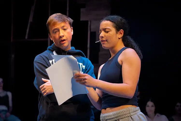 Josh Thone crosses his arms and looks down at a bundle of papers with Olivia Coleman. They are in an auditorium with other students looking on.