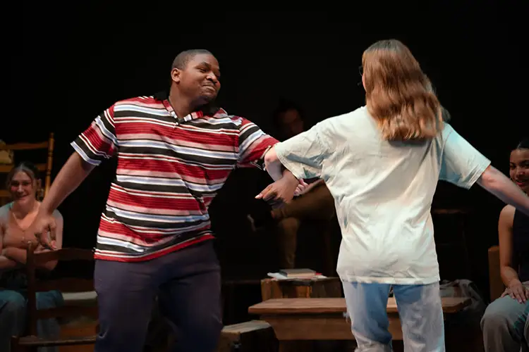 Sandders hooks are with a person, whose back is turned towards the camera) and smiles. They are in an auditorium with other students watching them.