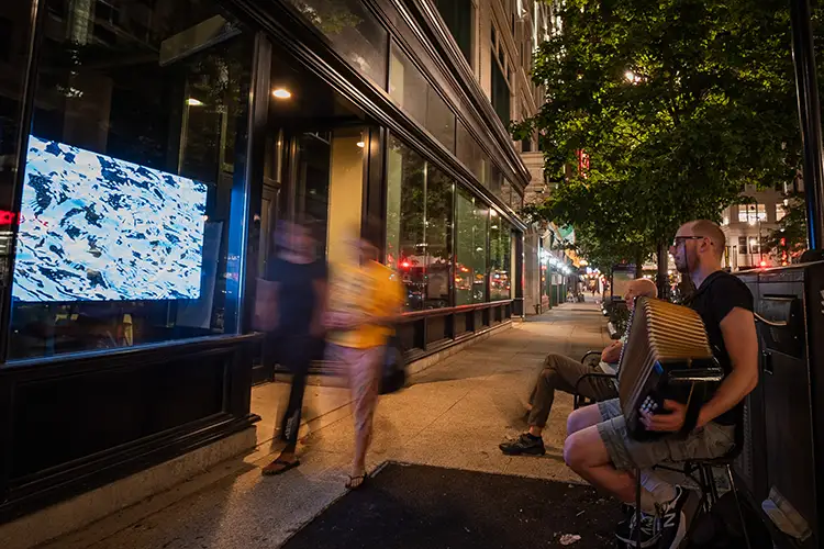 Two people walk down a city street. They are blurred. A man playing the accordian watches them go by.