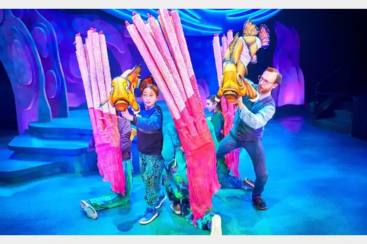 Performers on a vibrant stage hold large fish puppets among tall pink coral props in a colorful underwater-themed set. Performers on a vibrant stage hold large fish puppets among tall pink coral props in a colorful underwater-themed set.
