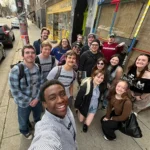 A group of people gathered on a sidewalk for a wide selfie, standing in front of storefronts and a building with colorful wooden boards.