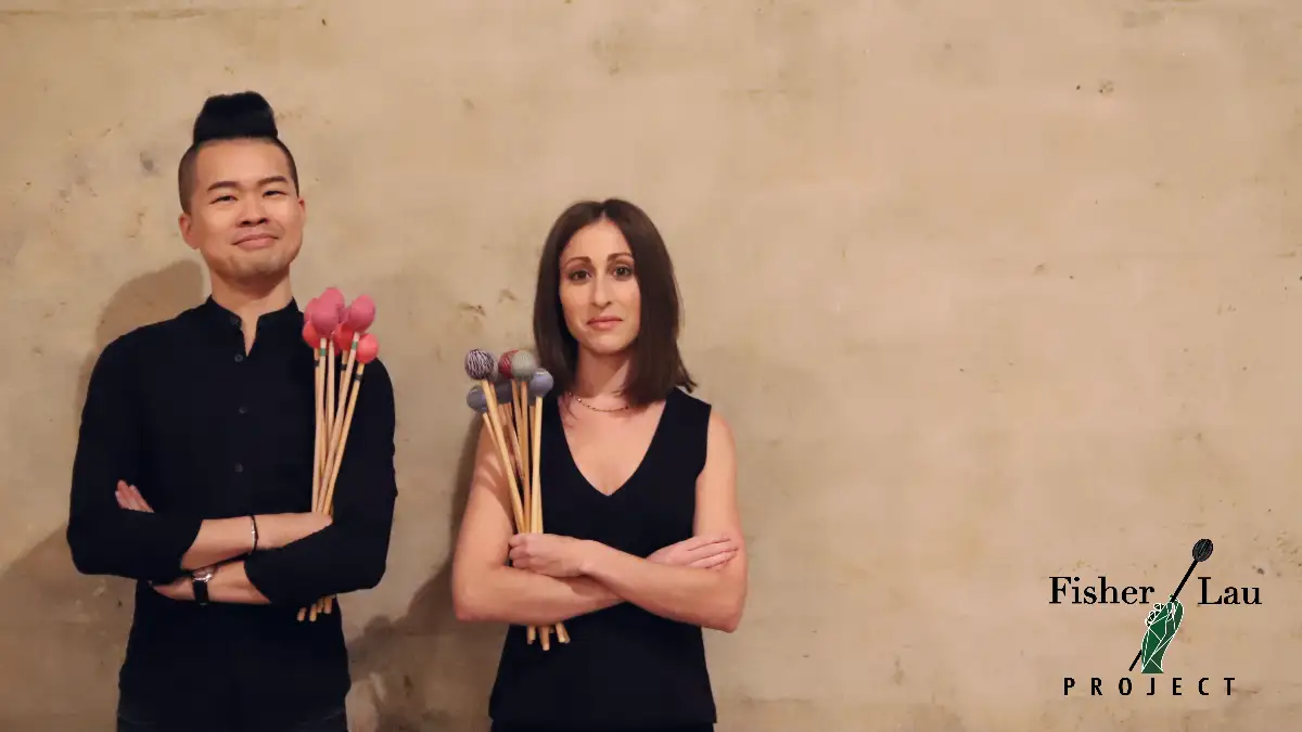 Two people standing against a wall holding multiple percussion mallets, with a logo beside them.