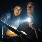 A man holds a flashlight pointed into darkness while a woman peers over his shoulder in the same direction.