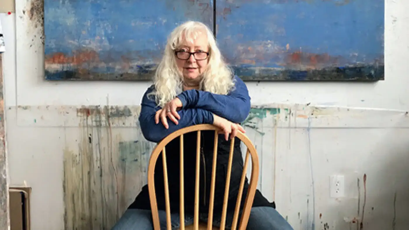 UWM Professor Allison B. Cooke seated backward on a wooden chair in front of a large abstract blue painting.