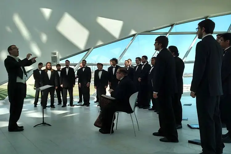 Choir performs in a sunlit modern gallery as a conductor leads singers before expansive windows overlooking the water. Choir performs in a sunlit modern gallery as a conductor leads singers before expansive windows overlooking the water.
