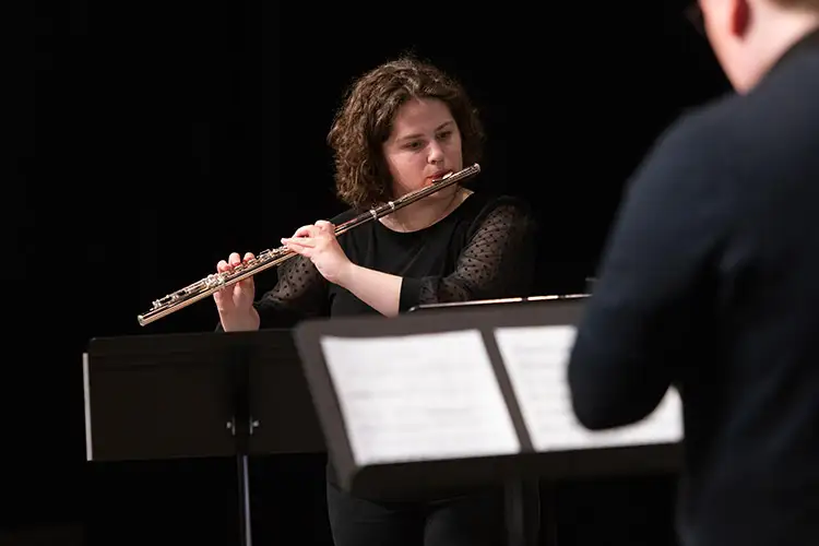 Person playing a flute during a music performance with sheet music stands in the foreground.