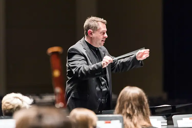 Conductor Dr. Scott Corley is seen holding a baton as he leads musicians during a performance.