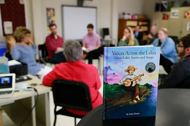 Music History and Literature BFA Book titled ‘Voices Across the Lakes’ on a table with a group discussion in the background.