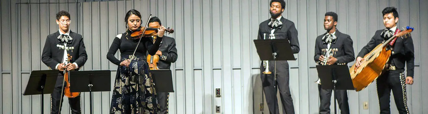 Mariachi ensemble performing on stage with violins, trumpet, and guitar in traditional attire.