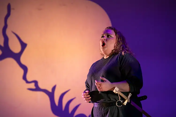 Music History and Literature MM Opera singer Scotlyn Haverkorn on stage in dark costume with a sword at her side and a large moon backdrop and tree silhouette.