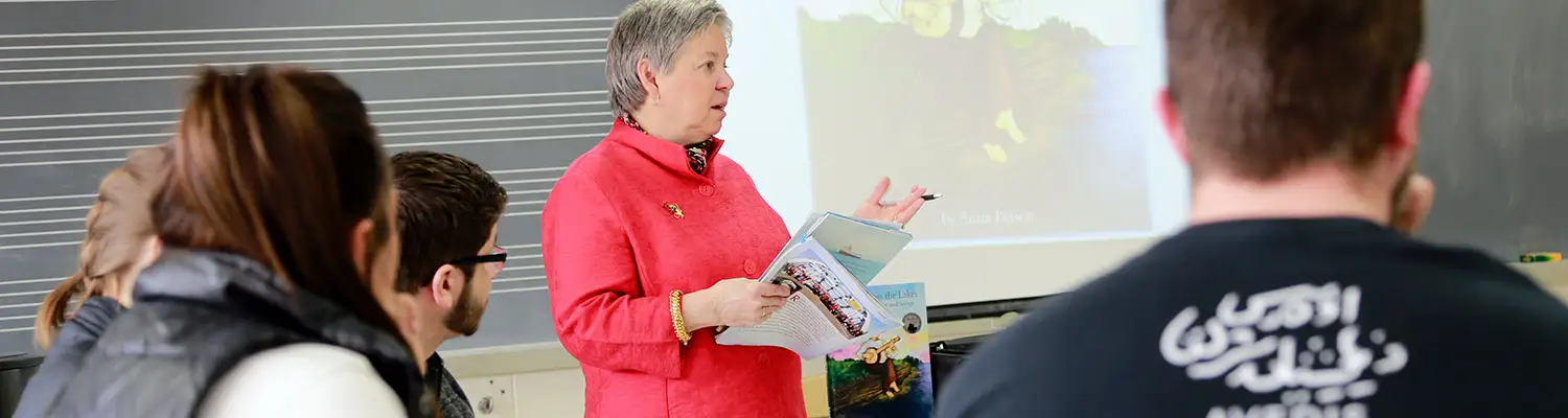 Music librarian instructor holding books and speaking to a group with a music staff board and projector.