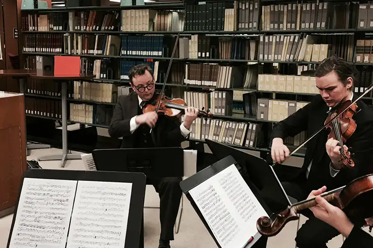 Music Library and Information Science MM Musicians performing a string ensemble in a library setting with sheet music and bookshelves in view.