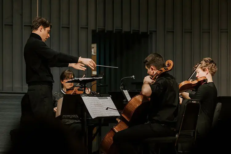 Conductor leading a small ensemble of string musicians performing with sheet music on stands.