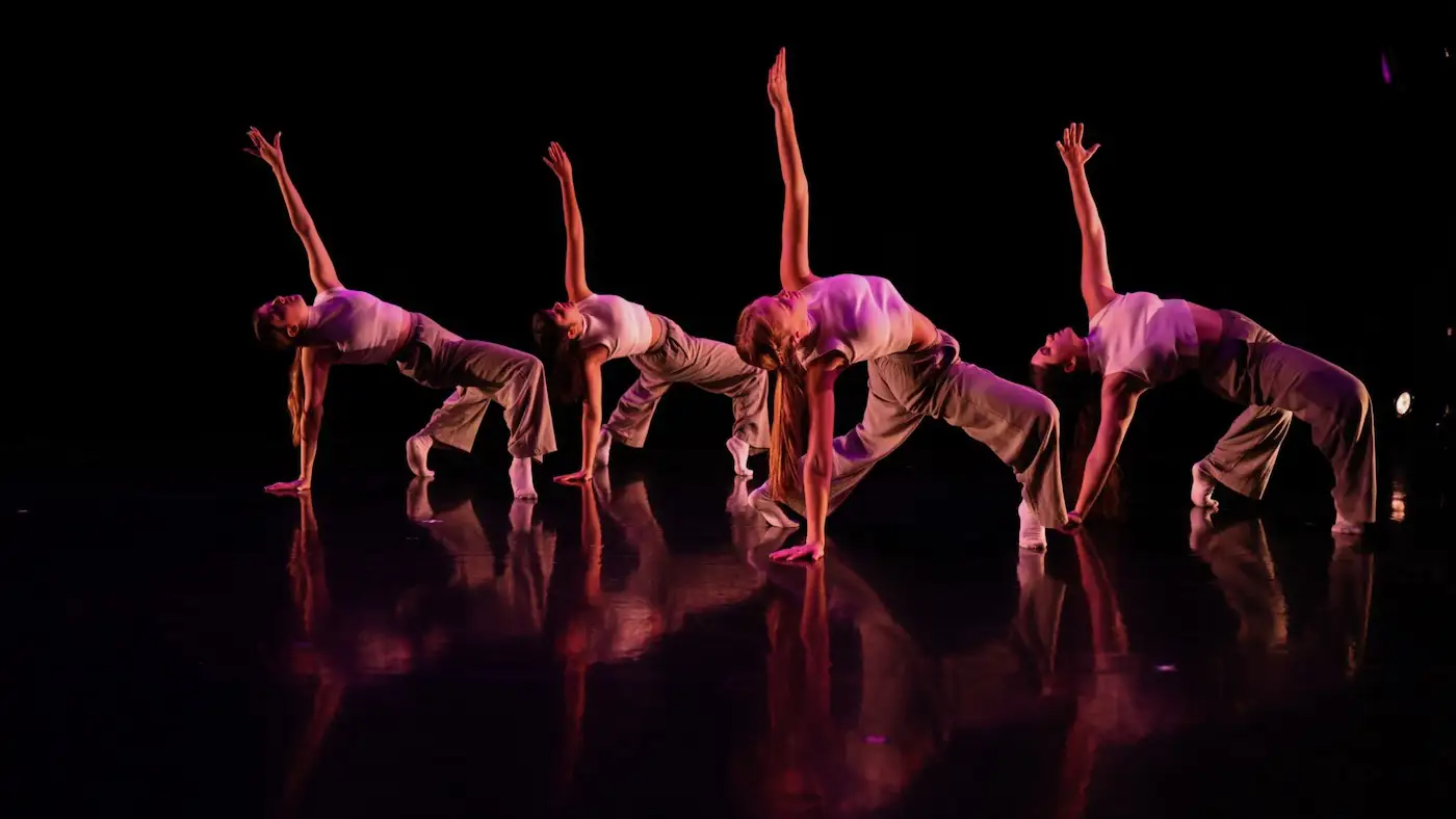 Five dancers in a line arch backward with one arm raised under magenta stage lighting.