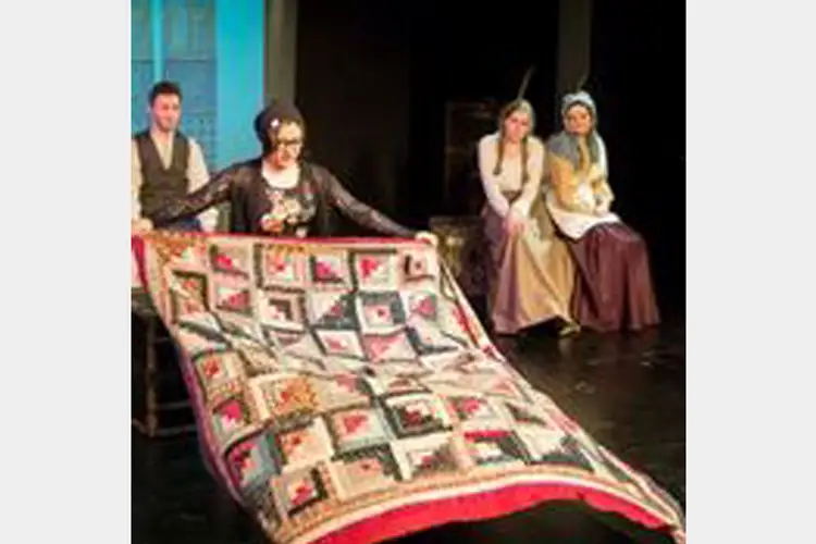 Stage scene with one performer holding a large patterned quilt in the foreground while three costumed performers sit behind on chairs.
