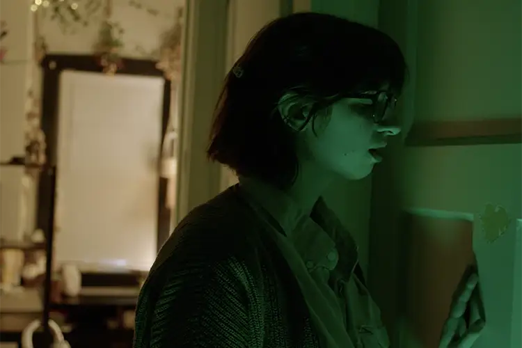 A young woman stands next to a closed door and is lit by green light A young woman stands next to a closed door and is lit by green light