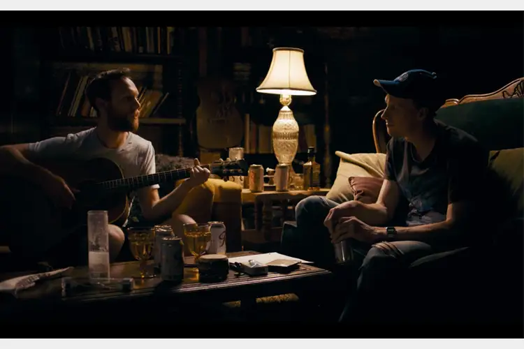 Two people sit in a dim, cozy room; one plays acoustic guitar while the other listens, with drinks and a lit lamp on a cluttered table. Two people sit in a dim, cozy room; one plays acoustic guitar while the other listens, with drinks and a lit lamp on a cluttered table.