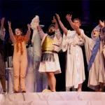 Actors on stage in costumes raising their hands in the air and singing.