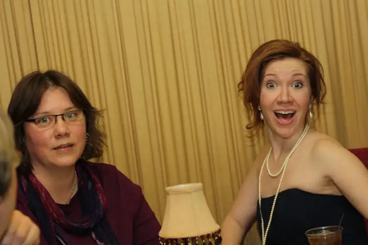 Two people seated at a table indoors, one wearing glasses and scarf, the other in a strapless dress with necklace, both facing the camera. Two people seated at a table indoors, one wearing glasses and scarf, the other in a strapless dress with necklace, both facing the camera.