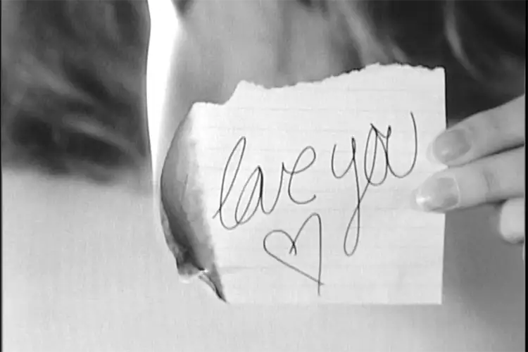Hand holding a small burning piece of paper with the handwritten words "love you" and a heart.