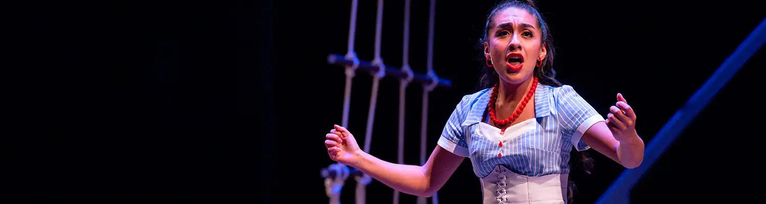 Stage performer in a striped costume sings under blue theater lighting beside rigging onstage.