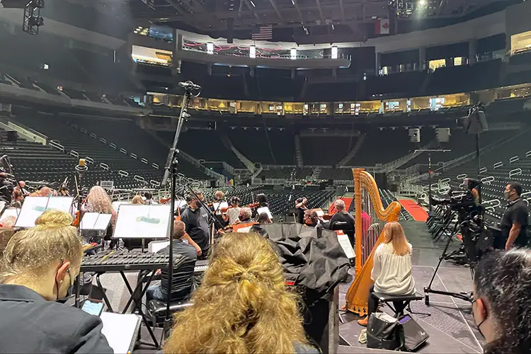 Orchestra rehearses on the floor of a large arena, with a harp, music stands, and empty seating visible throughout the venue. Orchestra rehearses on the floor of a large arena, with a harp, music stands, and empty seating visible throughout the venue.